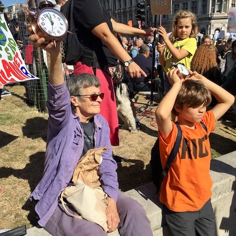 黛娜坐在头上，头上戴着一个时钟，显示已经快十二点了。 在她周围，一群人正忙着抗议。